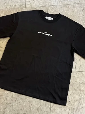 Svart Maison Margiela tshirt - Svart t-shirt från Maison Margiela med vit logotyptryck på bröstet i storlek M, T-shirten är i toppskick och fraktas inom 24h✅