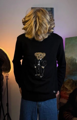 Svart stickad tröja med björn från Ralph Lauren - Svart stickad tröja från Ralph Lauren med en broderad björn i kostym på bröstet. Tröjan har rund hals, ribbade muddar och RL-broderi vid nederkanten. Perfekt för dig som gillar coola detaljer och klassisk streetstil.