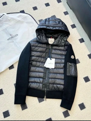 Svart pufferjacka från Moncler - Snygg svart pufferjacka från Moncler med huva och ribbstickade ärmar. Jackan har quiltad framsida, dragkedja och en praktisk ficka på ärmen. Materialmix av polyester och stickad ull ger både värme och stil. Perfekt för dig som vill ha en cool look. Storleken är M men passar även S 