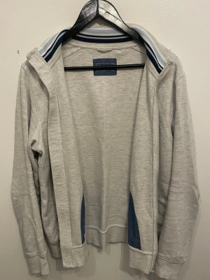 Ljusgrå hoodie med dragkedja - Ljusgrå hoodie med dragkedja och huva. Insidan av kragen har snygga blå och vita ränder, och det finns blå detaljer vid sidorna. Tröjan har långa ärmar och ribbade muddar. Perfekt för chill dagar eller när du vill ha en sportig look.