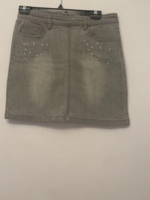 Grå kortkjol med pärlor från Soaked in luxury  - Har köpa den förlänge sedan men inte använt alls. Snygg grå kortkjol från Soaked med dekorativa pärlor vid fickorna framtill. Kjolen har klassisk jeanslook med bälteshällor, fickor och dragkedja bak. Perfekt för dig som gillar stilrena detaljer och vill ha en kjol som sticker ut lite extra. Passa på!!