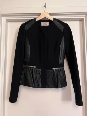 Zara Svart ullkavaj med skinndetaljer - Svart peplum ullkavaj från Zara med detaljer i skinnimitation på axlar, sidor och nedtill. Jackan har långa armar, guldfärgade dragkedjor och knappar framtill. 53% ull, så den är varm och skön. Mycket bra skick men har defekt i runt kragen (pga imitationslädret har skavts upp). Sälj på flera sidor, först till kvarn. 