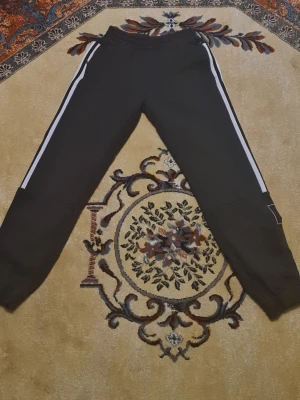 Svarta Adidas mjukisbyxor med vita ränder - Säljer ett par svarta mjukisbyxor från Adidas med klassiska vita ränder längs sidorna och elastisk midja. Byxorna har en patch med Adidas-logga på benet och är perfekta för chill eller träning. Tillverkade i mjukt material för maximal komfort.