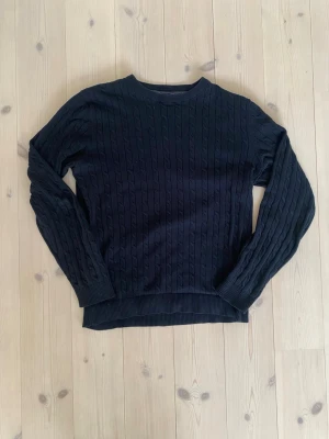 Svart kabelstickad tröja Jack & Jones - Kabelstickad svart tröja från Jack & Jones i storlek L. Klassisk modell med rund hals och ribbade muddar vid ärmslut och nederkant. Perfekt för lager på lager under höst och vinter. Snygg struktur som ger en stilren vibe.