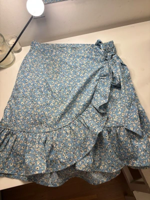 Blommig blå omlottkjol med volang - Supersöt blå omlottkjol med vitt blommönster och volangkant. Kjolen har en knytning i sidan som ger en extra detalj och flowig känsla. Perfekt för dig som gillar en feminin och somrig stil. Från Only i storlek S