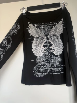Svart långärmad topp med vingar - Cool svart långärmad topp med stora silvriga vingar och detaljerat tryck framtill och på ärmarna. Toppen har en rundad halsringning och är figurnära i modellen. Materialet känns mjukt och stretchigt, 