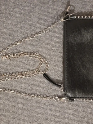 Svart axelväska med kedjerem - Säljer en svart axelväska i skinnimitation med silverfärgad kedjerem och dragkedja. Väskan har en stilren look och små nitar längs kanten för extra edge. Perfekt för dig som gillar minimalistisk men ändå cool stil.