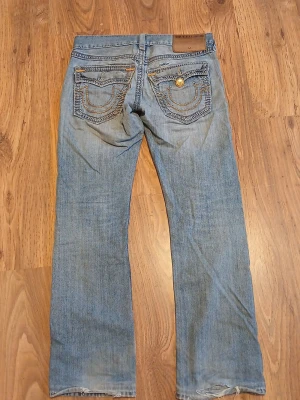 Blå True religion jeans  - Säljer ett par blå jeans från True Religion med snygga slitningar framtill och broderade bakfickor med kontrastsömmar och guldfärgade detaljer. Klassisk femficksmodell med raka ben och låg midja. Perfekta för dig som gillar en avslappnad och cool stil. Priset går att förhandlas då det fattas en knapp på bakfickan och det finns ett hål vid högra bakfickan. 