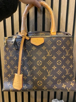  handväska brun  - Säljer en klassisk handväska i brunt och beige. Väskan har två robusta handtag, dragkedja upptill och flera fack inuti. Tillverkad i tåligt skinn med detaljer i ljusbrunt och insida i rött tyg. Perfekt för dig som gillar lyxig och tidlös stil.