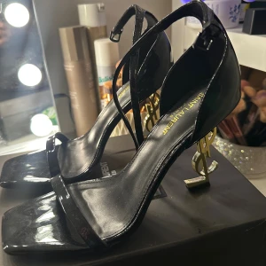 Svarta Saint Laurent klackar med logga - Säljer ett par svarta Saint Laurent klackar i lackat skinn med ikonisk YSL-logga i guld som klack. Skorna har smal rem över tårna, fyrkantig tå och justerbar ankelrem. Perfekta för dig som vill sticka ut med en lyxig och edgy vibe. Använd bara en gång