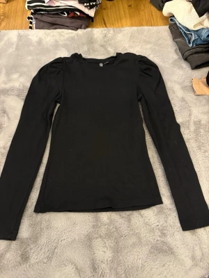 Svart ribbad topp från Pieces - Säljer en svart ribbad topp från Pieces i storlek S. Toppen har puffade axlar och långa ärmar vilket ger en trendig siluett. Materialet är mjukt och stretchigt, perfekt för att styla med jeans eller kjol. Enkel rund halsringning och figurnära passform.