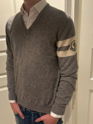 Grå Moncler stickad tröja v-ringad - Säljer en grå stickad tröja från Moncler med v-ringning och långa ärmar. Tröjan har två vita ränder på ena ärmen och Moncler-logga. Perfekt till skjorta under för en clean och stilren look.