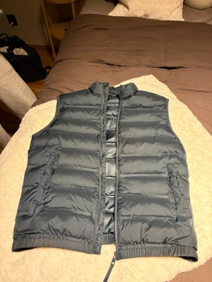 Mörkblå dunväst från Zara - Säljer en mörkblå dunväst från Zara med hög krage och dragkedja framtill. Västen har två fickor med lock på sidorna och quiltad design som ger en snygg och puffig look. Perfekt att slänga över en hoodie eller tröja för extra värme.