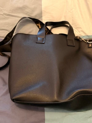 Svart axelväska från Even&Odd - Snygg svart axelväska från Even&Odd i strukturerat skinnimitation. Väskan har både handtag och en bred axelrem med mönsterdetaljer. Perfekt storlek för att få plats med allt du behöver till vardagen.