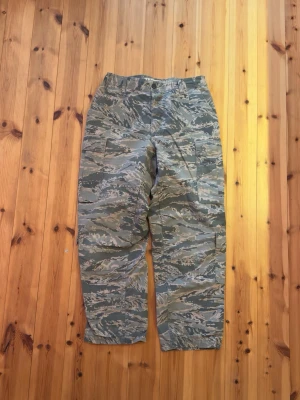 US Air Force camouflage cargobyxor - Säljer ett par feta cargobyxor med camouflage-mönster, tillverkade för U.S. Air Force. Byxorna har flera stora fickor på sidorna och bak, klassisk knappstängning och är gjorda i slitstarkt bomullsmaterial. Perfekta för dig som gillar utility-vibe och militärstil.