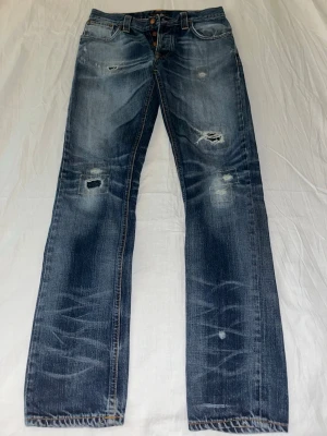 Nudie Jeans med slitningar - Säljer ett par blå Nudie jeans med slitningar och lagade detaljer framtill. Jeansen har raka ben, klassisk femficksmodell och knappar i gylfen. Snyggt tvättade med kontrastsömmar och bakfickor med broderad detalj. Storlek: 30/34