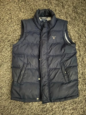 Mörkblå dunväst från Gant - Snygg mörkblå dunväst från Gant med hög krage och två fickor framtill. Västen har ett randigt innerfoder i vitt och blått samt tryckknappar med Gant-logga. Perfekt för lager-på-lager och klassisk stil. Ett litet hål på innersidan men man märker knappt.