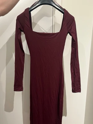 Vinröd långärmad klänning Bershka - Vinröd långärmad klänning från Bershka i storlek S. Klänningen har en snygg, fyrkantig urringning och är figurnära med lång siluett. Tillverkad i mjukt stretchigt material som sitter skönt på kroppen. Perfekt för dig som gillar minimalistisk och stilren look.