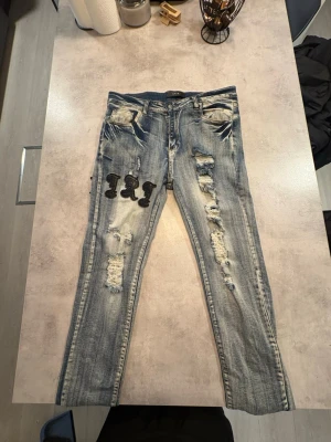 Amiri jeans - Säljer ett par blå jeans från Amiri med slitningar och patchade bokstäver på både fram- och baksida. Jeansen har en skinny passform, klassisk femficksdesign och coola detaljer som ger en edgy vibe. Materialet är jeans med tydliga slitningar och broderade detaljer.