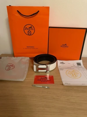 vit läderbälte med H-spänne - Säljer ett exklusivt bälte i vitt läder med den ikoniska H-spännet i silver. Bältet är stilrent och har snygga sömmar längs kanterna. Kommer med originalpåse, box, certifikat och kvitto. Perfekt accessoar för dig som vill ha något lyxigt och tidlöst.