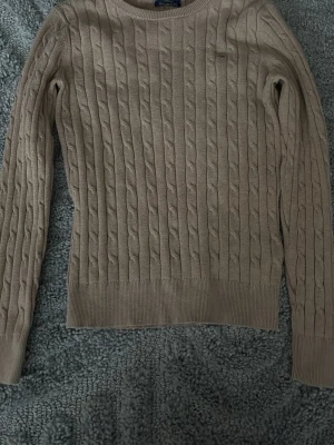 Beige kabelstickad tröja från Gant - Säljer en beige kabelstickad tröja från Gant i stretchig bomull. Klassisk rund halsringning och diskret Gant-logo broderad på bröstet. Perfekt till lager på lager och funkar året runt. Långärmad och med ribbade muddar för en snygg passform. 