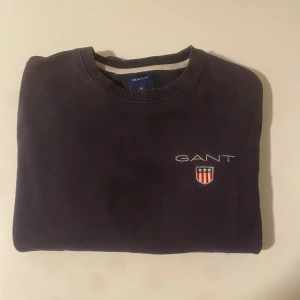 Mörkblå sweatshirt från GANT - Säljer en mörkblå sweatshirt från GANT med broderad logga och sköld på bröstet. Klassisk rund halsringning och ribbade muddar vid ärmar och nederkant. Perfekt till chill dagar och enkel att matcha med jeans eller sweatpants.