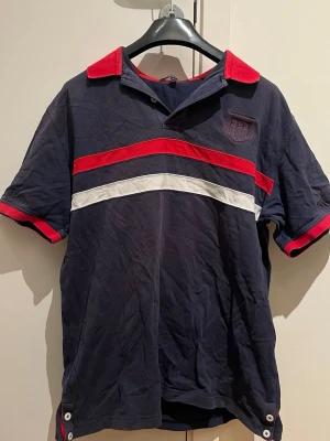 Marinblå randig pikétröja från Gant - Säljer en marinblå pikétröja från Gant i storlek XL. Tröjan har röda och vita ränder över bröstet, röd krage och röda detaljer vid ärmsluten. På bröstet finns ett broderat märke med stjärnor. Klassisk piké i bomull med knappar vid sidan.