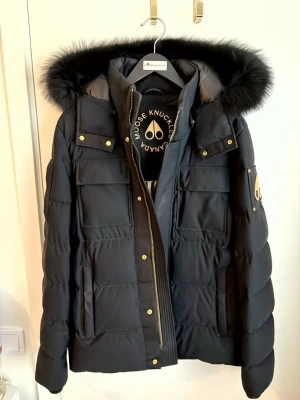 Svart  Moose Knuckles Jacka  - Säljer en svart pufferjacka från Moose Knuckles med guldfärgade detaljer och stor luva med fluffig päls. Jackan har flera fickor, dragkedja och tryckknappar. Klassisk logga på ärmen och riktigt varm för kalla dagar. Nypris 13.500