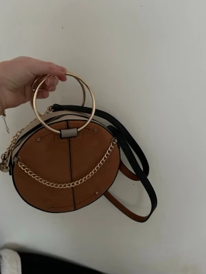 Rund brun axelväska med guldkedja - Säljer en rund axelväska i brun mockaimitation med svarta detaljer och guldfärgade metalldelar. Väskan har en snygg guldkedja framtill, metallhandtag och dragkedja upptill. Insidan är svart och väskan har även en beige ovansida. Helt oanvänd 
