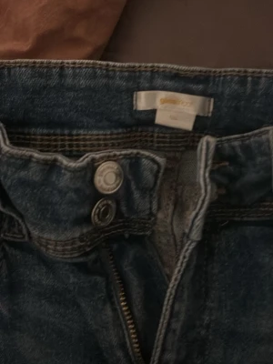 Blå jeans från Gina Tricot straight fit - Säljer ett par klassiska blå jeans från Gina Tricot i rak modell. Jeansen har normal midja, fem fickor och snygg slitning framtill. Materialet är robust denim med dragkedja och dubbla knappar i midjan. Perfekta till en avslappnad look.