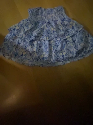 Blommig volangkjol i blått och vitt - Supersöt kort kjol med blått och vitt blommönster och bred smock i midjan. Kjolen har dubbla volanger som ger en riktigt flowy känsla och är perfekt för varma dagar. Lätt och luftig, passar perfekt till sneakers eller sandaler.