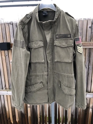 Olivgrön parkajacka från Smog XXL - Säljer en olivgrön parkajacka från Smog i storlek XXL. Jackan har militärstil med stora fickor, patchar som U.S. Defence och amerikansk flagga samt trycket 'ARMY' på ryggen. Tillverkad i bomull och har dragkedja och knappar framtill. OBS Liten storlek. Är mer som XL.