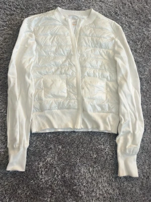 Vit quiltad jacka från Gina Tricot - Säljer en vit quiltad jacka från Gina Tricot i storlek 170. Jackan har dragkedja framtill, två fickor och ribbade muddar vid ärmslut och midja. Materialmix med glansig polyester på framsidan och mjuka stickade ärmar. Perfekt för dig som gillar en clean och fräsch look.