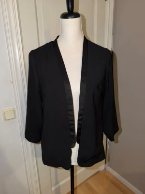 Svart kavaj med satinkant - Stilren svart kavaj med trekvartsärm och snygg satinkant längs öppningen. Rak passform som ger en clean look och funkar till många olika outfits. Perfekt för dig som vill ha en enkel men ändå elegant jacka i garderoben.