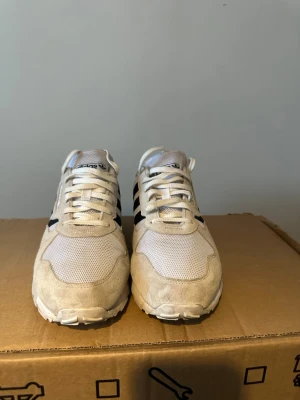 Vita Adidas sneakers med mockadetaljer - Säljer ett par klassiska Adidas sneakers i vitt med svarta ränder. Skorna har ovandel i mesh och mocka, samt snörning framtill. Ikoniska Adidas-loggor på plös och häl. Perfekta för dig som gillar retrostil och bekväma vardagsskor.