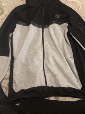 En svart/grå Nike tech - Nike zip-hoodie/jacka i svart och grått. Sportig modell med dragkedja framtill och Nike-logga på bröstet. Normal passform, mjukt material och bekväm att ha på sig både till vardags och träning. Storlek L. I fint skick.