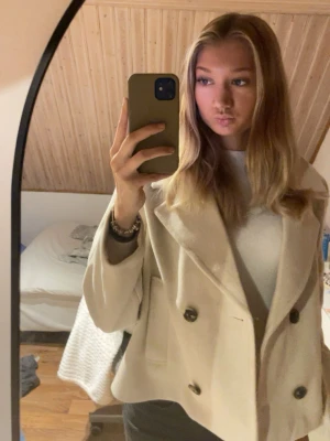 Beige skepparkavaj med stora knappar - Säljer en snygg beige skepparkavaj med bred krage och dubbla rader stora svarta knappar. Jackan har en klassisk, lite boxig passform och är perfekt för dig som vill ha en stilren look. Materialet känns mjukt och värmande, och den är enkel att matcha till olika outfits.