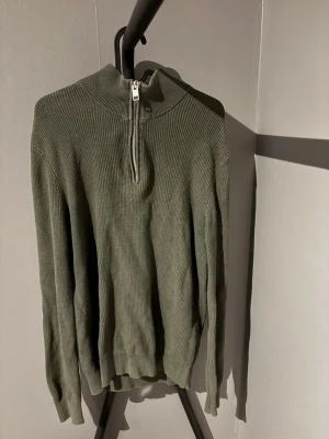 Olivgrön ribbad tröja med half zip - Säljer en olivgrön ribbad tröja med half zip och hög krage. Tröjan är långärmad och har en normal passform. Perfekt för dig som gillar stilrena och bekväma plagg till vardags.