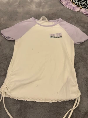 Ljuslila och vit topp från H&M - Säljer en söt topp från H&M med ljuslila raglanärmar och vit kropp. Tryck på bröstet med texten 'GOOD VIBES EVERYDAY'. Toppen har korta ärmar, ribbade kanter och snörning i sidorna för en extra detalj.