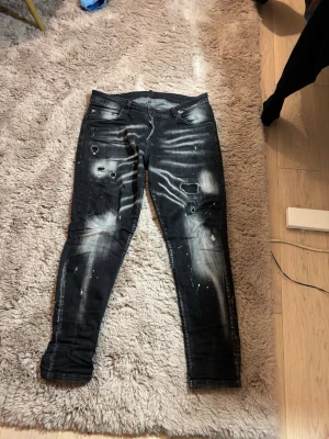 Svarta slimfit  jeans från Dsquared2 - Säljer ett par svarta skinny jeans från Dsquared2 med slitningar, färgstänk och coola detaljer. Jeansen har en tvättad look med vita partier och trasiga effekter på benen. Klassisk femficksmodell och låg midja. Perfekta för dig som vill sticka ut.