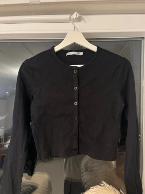 Svart croppad tröja från Lager 157 - Svart croppad tröja från 157 med rund halsringning och långa ärmar. Tröjan har ribbad struktur och knäpps med svarta knappar framtill. Perfekt till alla olika looks.