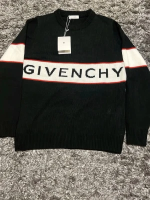 Svart stickad tröja från Givenchy - Säljer en svart stickad tröja från Givenchy med vit och röd rand över bröstet och armarna. Stor Givenchy-logga framtill. Tröjan har rund halsringning och långa ärmar. Perfekt för dig som vill ha en stilren och exklusiv look.