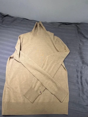 Beige polotröja Tommy Hilfiger ull - Snygg beige polotröja från Tommy Hilfiger i lyxig ull. Klassisk och stilren design med ribbade muddar och diskret logga på ärmen. Perfekt för dig som vill ha en clean och tidlös look till höst och vinter.