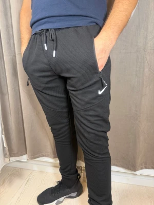Svarta mjukisbyxor från Nike - Säljer ett par svarta mjukisbyxor från Nike med vit logga på låret. Byxorna har dragsko i midjan, dragkedjor vid fickorna och är tillverkade i ett ribbat syntetmaterial. Passformen är slim och de är perfekta för träning eller chill.