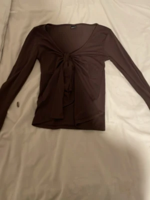 Brun långärmad topp med knyt - Säljer en brun långärmad topp med snygg knytning framtill och djup v-ringning. Tunn och stretchig, perfekt att styla med jeans eller kjol. Enkel men trendig design som ger en cool vibe.