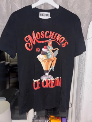 Svart Moschino t-shirt med glassmotiv - Cool svart t-shirt från Moschino med färgglatt retroinspirerat glassmotiv och stor text 'Moschino's Ice Cream' på framsidan. Klassisk passform och rund halsringning. Perfekt statement-plagg för dig som gillar unika prints.