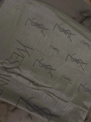 Grå väst med YSL-logga - Säljer en grå väst från Yves Saint Laurent med ikoniska YSL-loggor invävda över hela plagget. Västen har en stilren look med diskret mönster och är tillverkad i ett mjukt material som känns lyxigt mot huden. Perfekt för dig som gillar designerplagg med subtila detaljer.