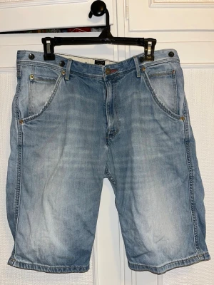 Ljusblå jeansshorts från Lee - Säljer ett par ljusblå jeansshorts från Lee med klassisk femficksdesign och Lee-lapp bak i midjan. Shortsen har normal passform, bälteshällor och dragkedja med knapp. Perfekta för sommaren och riktigt chill stil. Litet hål längst ner i mitten på baksidan men syns inte när man har på shortsen, lätt att laga för den händiga. 