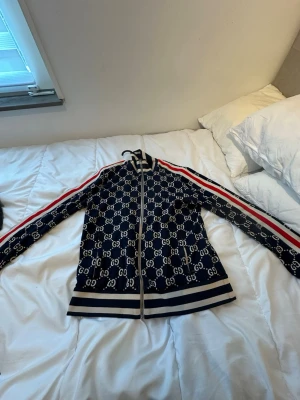 Gucci blå kofta med GG-mönster - Säljer en ikonisk marinblå kofta från Gucci med helvitt GG-mönster, röda och vita ränder längs ärmarna samt ribbade muddar och nederkant. Dragkedja framtill och klassisk sportig look. Perfekt för dig som vill sticka ut med lyxig streetstyle. Byxa finns till, ( ett sett) vid fler bilder hör av dig 
