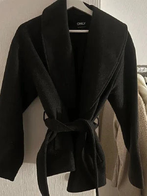 ONLY kappa - Snygg svart jacka från ONLY med bred krage och knytskärp i midjan. Jackan har en omlottmodell och är tillverkad i ett mjukt material. Perfekt till vår och höst